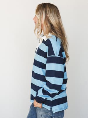 A side-profile shot of a model in a bold navy and sky blue striped rugby top with a white collar, paired with denim — easy, relaxed, and the kind of top that makes Saturday errands feel like a personality.
