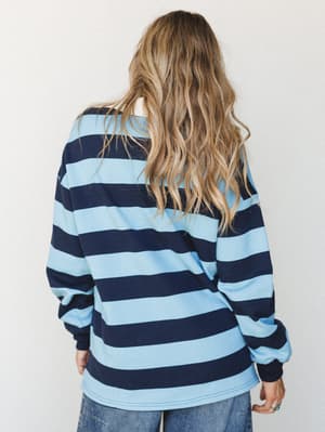 Shot from the back, a model with loose wavy hair wears an oversized long-sleeve rugby-stripe top in sky blue and navy, paired with wide-leg denim — casual, lived-in, and effortlessly cool.