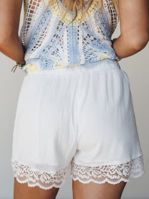 A close-up back view of white gauze shorts with a wide lace hem trim, paired with a multicolor crochet top — light, airy, and effortlessly boho from waist down.