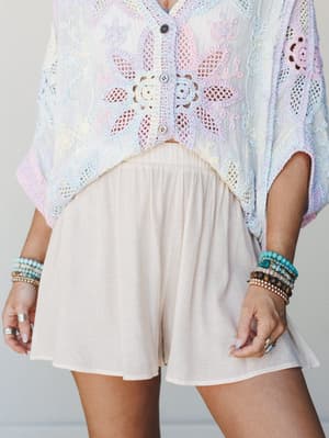 A dreamy mid-shot pairing cream flowy shorts with a pastel multicolor crochet top, styled with stacked boho bead bracelets — the kind of outfit that feels like a long weekend with nowhere to be.