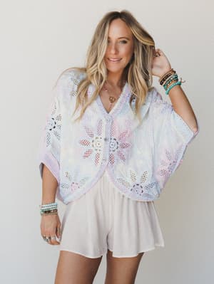 A flowy pastel crochet cardigan with wide bat-wing sleeves and floral cutwork layered over simple cream shorts — the kind of outfit that feels like a daydream you actually got dressed in.
