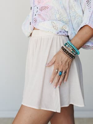 A close-up lifestyle shot showing a cream mini skirt paired with a pastel multicolor crochet top, styled with a stacked bracelet set and turquoise stone rings — soft, dreamy, and full of that effortless boho layering energy.