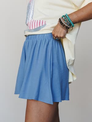 A close side-angle shot of a flowy dusty blue mini skort styled with a soft yellow tee and a striped embroidered bag, accessorized with a stack of beaded bracelets and a turquoise ring — easy, collected summer dressing.