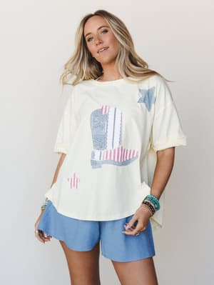 A breezy, lived-in look — an oversized cream graphic tee with a patchwork cowboy boot print paired with soft dusty blue shorts, styled with stacked beaded bracelets for that effortless, easygoing energy.