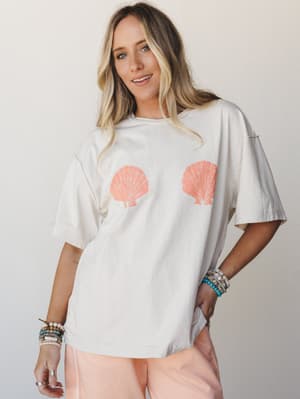An oversized cream tee with a playful double scallop shell graphic gets styled with blush shorts and a stack of beaded bracelets — casual, coastal, and the kind of thing you'd reach for on a beach-town morning.