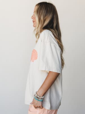 A side-profile shot of a model in an oversized cream graphic tee with a coral seashell print on the chest, paired with blush shorts and a stack of beaded bracelets — easy, sun-worn, and ready for wherever the summer takes you.