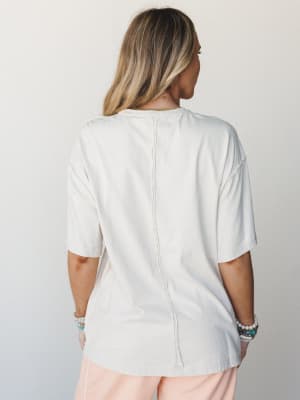 A back-view shot of a relaxed, oversized cream tee with a visible center back seam detail, styled with blush wide-leg pants and layered beaded bracelets — easy and lived-in in the best way.