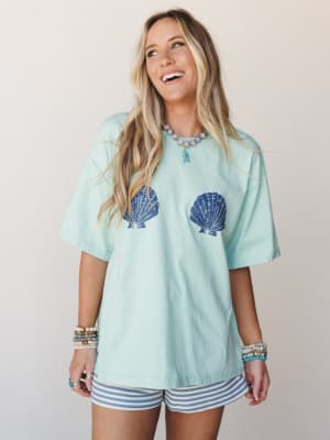 A smiling model wears an oversized mint tee with bold navy scallop shell graphics paired with blue and white striped shorts, layered bead bracelets, and a fish pendant necklace — easy, coastal, and genuinely happy.