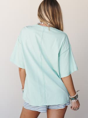 A breezy mint oversized tee shot from the back, styled with striped shorts and layered beaded bracelets — the kind of easy, sun-warmed outfit that feels like a slow Saturday morning.