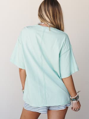 A breezy mint oversized tee shot from the back, styled with striped shorts and layered beaded bracelets — the kind of easy, sun-warmed outfit that feels like a slow Saturday morning.