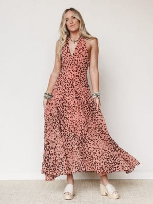 A flowy leopard-print halter maxi dress in warm dusty rose and brown swirls around the model as she stands against a soft white wall — styled with stacked boho bracelets and white espadrille platforms for that effortless 'going somewhere good tonight