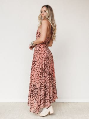 A flowy leopard-print maxi dress in dusty rose and brown photographed from a back-angle on a model with long wavy blonde hair — the open back and espadrille wedges give it that effortless warm-weather energy.