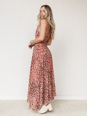 A flowy leopard-print maxi dress in dusty rose and brown photographed from a back-angle on a model with long wavy blonde hair — the open back and espadrille wedges give it that effortless warm-weather energy.