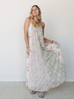 A flowy floral maxi dress in cream and sage panels catches mid-movement, styled with stacked bracelets and a layered necklace — the kind of dress that makes you want to find a reason to wander somewhere warm.
