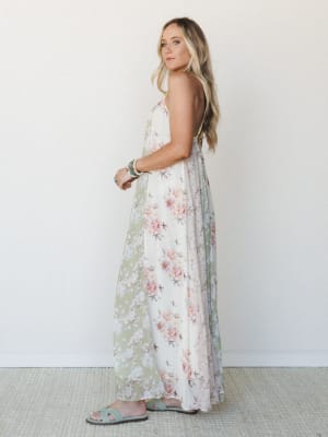 A flowy floral maxi dress in cream and sage green with soft pink roses, styled with slide sandals and stacked bracelets — the kind of dress that makes you want to find a garden party to walk into.