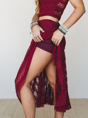 A rich burgundy lace two-piece set — cropped top and open-front maxi skirt layered over matching shorts — styled with stacked beaded bracelets and silver rings for a boho-romantic feel that reads festival-ready or date night without trying too hard.