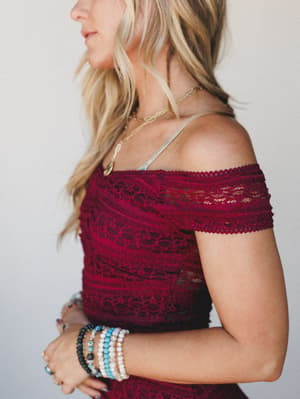 A close-up profile shot of a burgundy lace off-shoulder top, styled with layered beaded bracelets and a delicate gold necklace — romantic and a little undone in the best way.