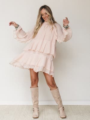 A flowy blush mini dress with tiered ruffles and dramatic long sleeves catches mid-twirl, styled with knee-high suede boots and stacked turquoise bracelets — the kind of outfit that makes you want to spin in a parking lot.