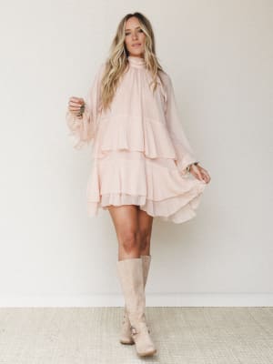 A flowy blush mini dress with tiered ruffles and long sheer sleeves styled with tall suede boots — soft, romantic, and the kind of outfit that moves when you do.