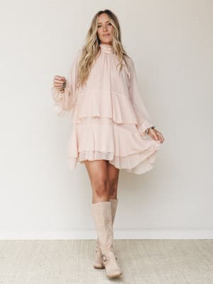 A flowy blush mini dress with tiered ruffles and long sheer sleeves styled with tall suede boots — soft, romantic, and the kind of outfit that moves when you do.
