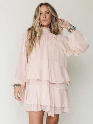A flowy blush mini dress with cascading tiered ruffles and dramatic bishop sleeves styled with stacked beaded bracelets — soft, dreamy, and made for someone who wants to feel like the main character at a bridal shower or garden party.