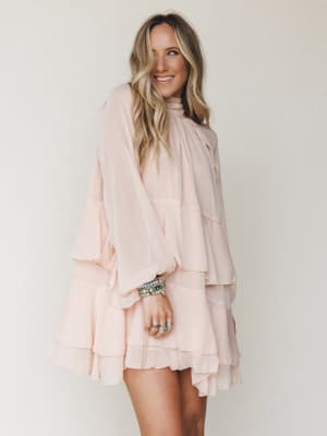 A flowy blush mini dress with tiered ruffles and dramatic bell sleeves styled against a soft neutral backdrop — the kind of dress that makes you want to twirl before you even put it on.