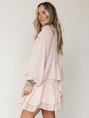 A flowy blush mini dress with tiered ruffle skirt and billowy long sleeves, styled with stacked boho bracelets and rings — soft, feminine, and effortlessly easy to wear.