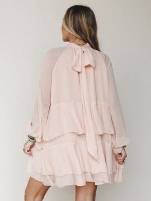 A flowy tiered mini dress in soft blush pink shot from the back, showing off a delicate bow tie neckline and billowy long sleeves — the kind of dress that moves with you and makes even a Tuesday feel like something.