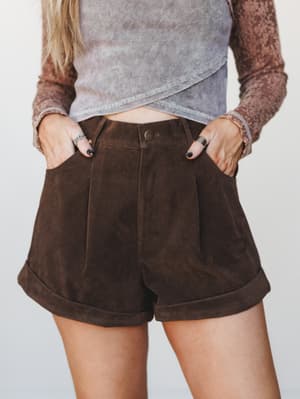 A close mid-shot of chocolate brown suede-look pleated shorts styled with a layered crossover ribbed crop top and a floral long-sleeve underneath — earthy, intentional, and very much the kind of outfit you'd put together on a good day.