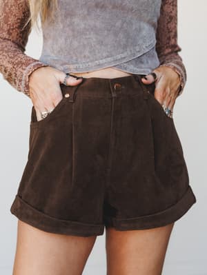Rich chocolate suede-look pleated shorts with cuffed hems styled with a layered rib crop top and floral lace overlay top — earthy, relaxed, and effortlessly put-together.