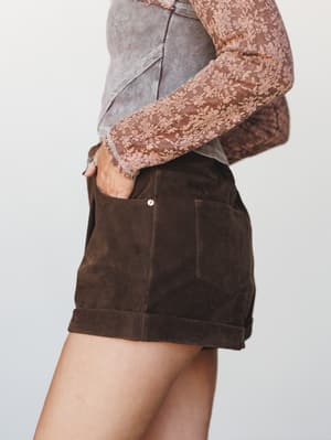 A close side-angle shot of rich chocolate suede shorts with cuffed hems, styled with a floral lace long-sleeve top and a cropped denim layer — earthy, vintage-feeling, and very much the kind of outfit you'd put together on a good day.