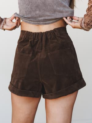 A close-up back view of rich chocolate corduroy high-waist shorts with cuffed hems and patch pockets, styled with a striped crop top and a floral-sleeved layer — earthy and effortlessly put-together.