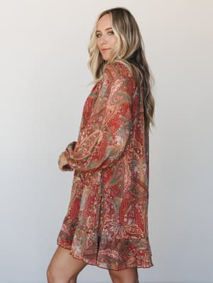 A flowy paisley mini dress in rich rust and sage, styled on a model against a clean light backdrop — the kind of dress that makes a Tuesday feel like a reason to go somewhere.