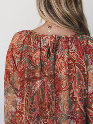 A close-up shot of the back neckline detail on a rich rust paisley dress — the tie closure and gathered yoke are front and center, with a delicate gold charm necklace peeking out above.