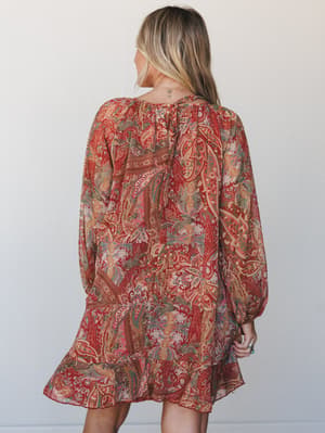 A flowy mini dress in a rich rust and multi-color paisley print, shot from the back to show the relaxed silhouette, billowy long sleeves, and ruffle hem — the kind of dress that feels like a vintage find but fits like it was made for you.