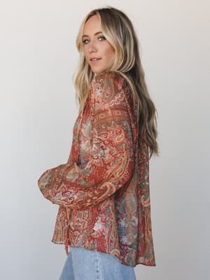 A paisley-printed sheer blouse in warm rust and sage tones styled with light wash denim — the kind of top that looks like it has a story, shot against a clean studio backdrop with a soft, natural feel.