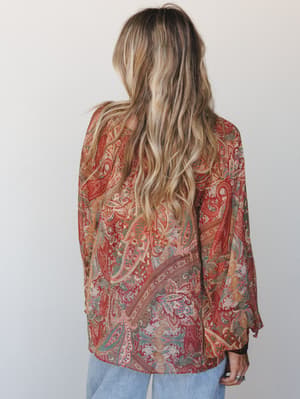 A back-facing model with loose wavy hair wears a flowy paisley blouse in warm rust, sage, and dusty rose tones, paired with light-wash jeans against a soft white studio backdrop — effortlessly boho and lived-in.
