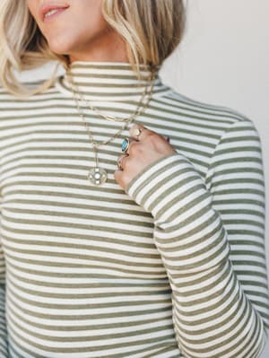A close-up shot of a sage-striped mock neck long sleeve top, styled with layered gold necklaces and stacked rings — the kind of effortless detail that makes a simple top feel like a whole mood.