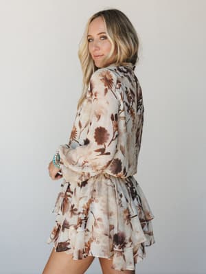 A flowy floral mini dress in cream and warm brown photographed from a three-quarter back angle — the tiered ruffle skirt and billowy long sleeves give it that effortless, romantic energy that looks just as good at brunch as it does at a summer weddin