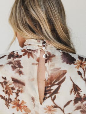 A close-up back detail shot of a floral blouse or dress in warm cream and earthy botanicals, highlighting a keyhole back opening secured with a fabric-covered button and a delicate gold pendant necklace peeking through.