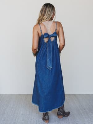 Woman wearing a blue denim dress with a lace back and bow details, standing against a white wall.