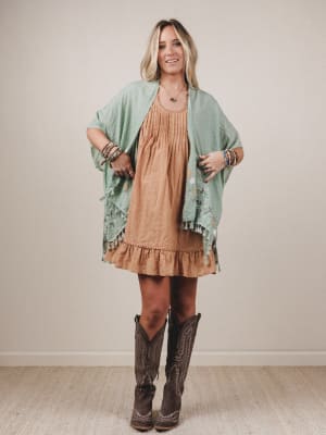 Woman wearing a tan dress with a green shawl and brown cowboy boots against a plain background