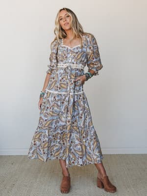 A flowy paisley midi dress in soft powder blue gets the full boho treatment — lace trim at the waist and neckline, puff sleeves, and a tiered skirt styled with brown clogs and stacked turquoise bracelets for that effortless, lived-in look.