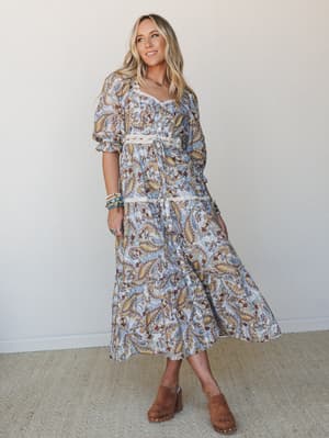 A paisley midi dress in powder blue with golden and burgundy tones, styled with brown clogs and stacked turquoise bracelets — the kind of outfit that looks equally at home at a farmers market or a backyard wedding.