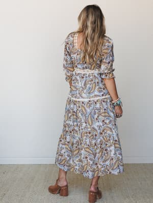 A back-facing shot of a tiered paisley midi dress in powder blue with lace trim detailing at the waist, styled with wood-platform clogs and layered turquoise bracelets — the kind of dress that makes a Tuesday feel like a road trip.