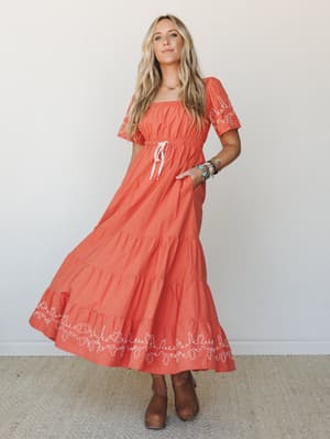 A tiered maxi dress in warm terracotta with white embroidery trim along the hem and sleeves, styled with stacked turquoise bracelets and tan clogs — the kind of dress that makes a Tuesday feel like something worth remembering.