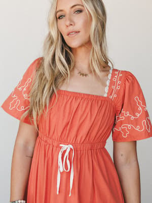 A warm terracotta dress with playful white embroidery on the sleeves and eyelet trim at the neckline — the kind of dress that makes a Saturday feel intentional without trying too hard.