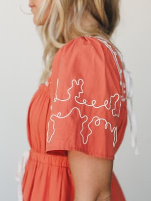 A close-up side shot of a terracotta dress with whimsical white embroidery along the sleeve, white ribbon tie at the shoulder, and a cinched elastic waist — warm, handcrafted, and quietly expressive.