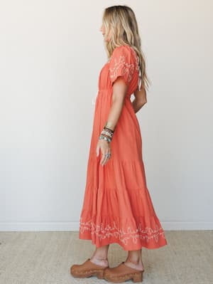 A warm terracotta midi dress with cream embroidery details and a tiered skirt, styled with stacked boho bracelets and tan clogs — the kind of dress that makes a Tuesday feel like a trip somewhere worth going.
