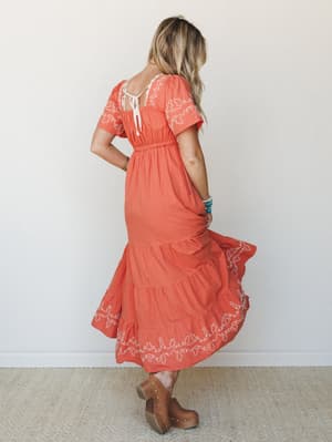 A model twirls away from camera in a warm terracotta midi dress with cream embroidery and a tiered skirt — the kind of dress that looks equally at home at a summer farmers market or a backyard wedding.